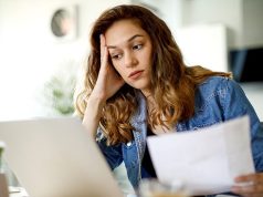 戴夫·拉姆齐告诉年轻美国人如何反击房屋所有权的“困境” A worried woman sits at a table reviewing her household budget and bills, appearing stressed about home finances.